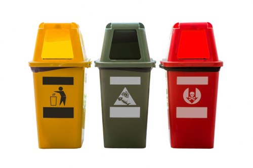 A photograph features three empty household waste bins placed side by side on a plain white background, each with a distinct colour and label for sorting different types of rubbish. The yellow bin on the left has a black icon of a person disposing of small waste into a container, indicating general waste; the green bin in the middle displays a white label and a diagram suggesting it is for organic or garden waste; the red bin on the right has a white label with a symbol of a biohazard or hazardous waste, indicating it is for dangerous or hazardous materials. Each bin has a matching lid, with the yellow bin's lid slightly open, revealing a smooth, plastic surface. The bins are made of durable plastic with textured surfaces to provide grip, and they are positioned on a clean, flat surface, likely outdoors or on a driveway, with no other objects or background details visible. The arrangement and design of the bins are consistent with waste disposal and rubbish management services offered by companies such as Commercial Waste Sidcup, serving local areas including Sidcup and the surrounding postcode regions, emphasizing efficient and organized rubbish collection solutions.