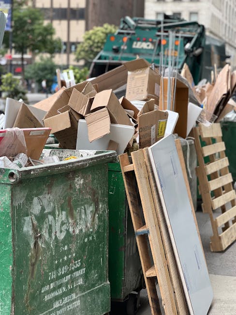 A collection of overflowing rubbish bins and scattered waste materials situated on a paved area near a parking lot. The central bin is a large, grey mixed waste container filled to capacity, with its 
